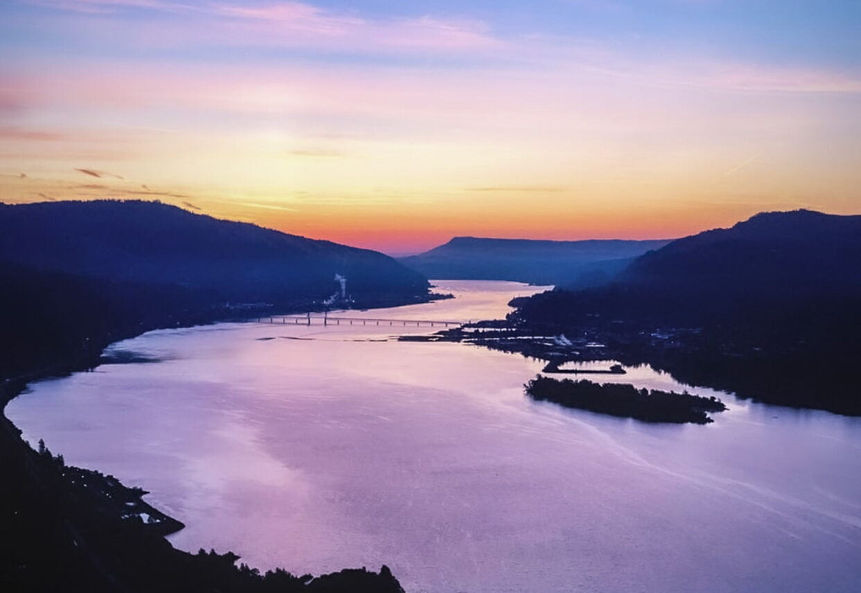 A wide, calm river at sunset reflecting the purple and peach of the sky. Blue hills surround the river.