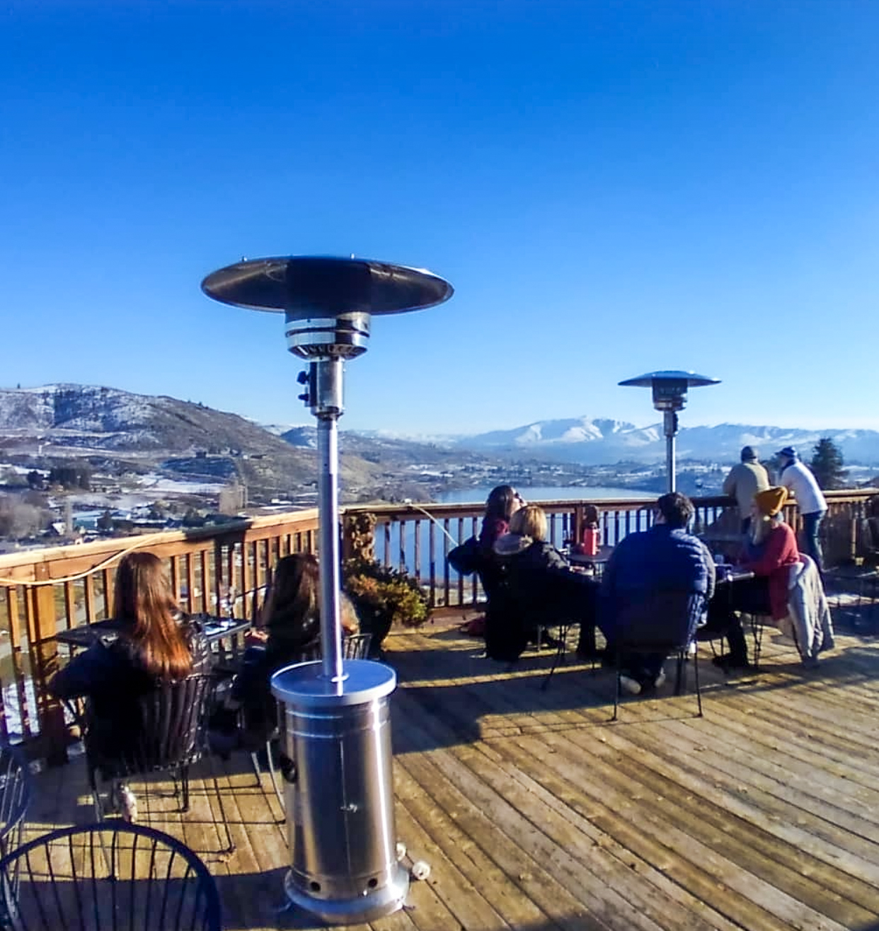 Deck with people at tables and heat lamps, overlooking a large lake. Snow-covered mountains are in the distance, with a bright blue winter sky.