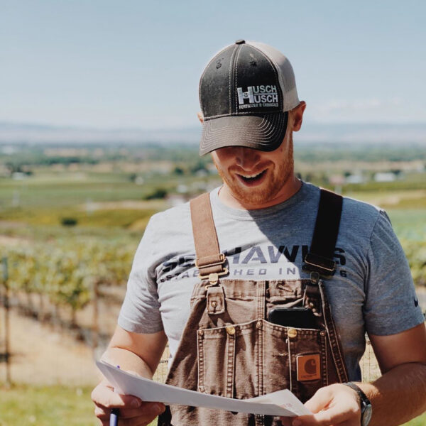 A man wearing overalls and a ballcap looks down at a paper in his hands as he speaks. Vineyards and a blue sky are behind him.