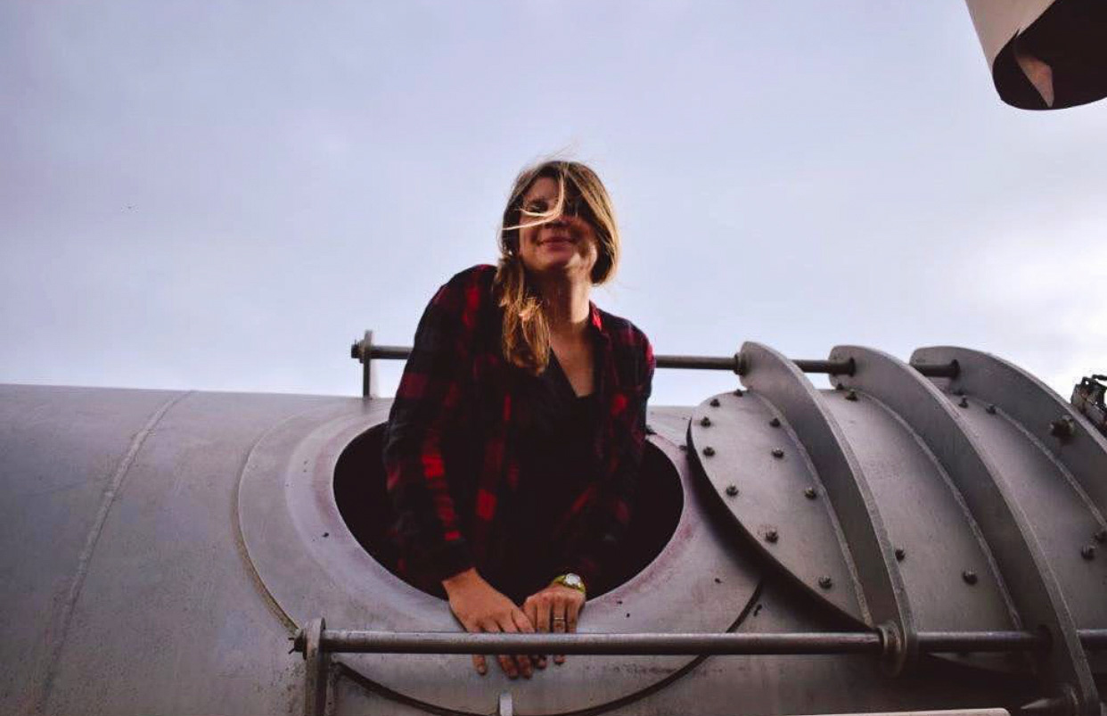 A woman in a red and black plaid shirt leans out the circular opening of a large metal vessel. She is smiling and her hair blows in the wind.