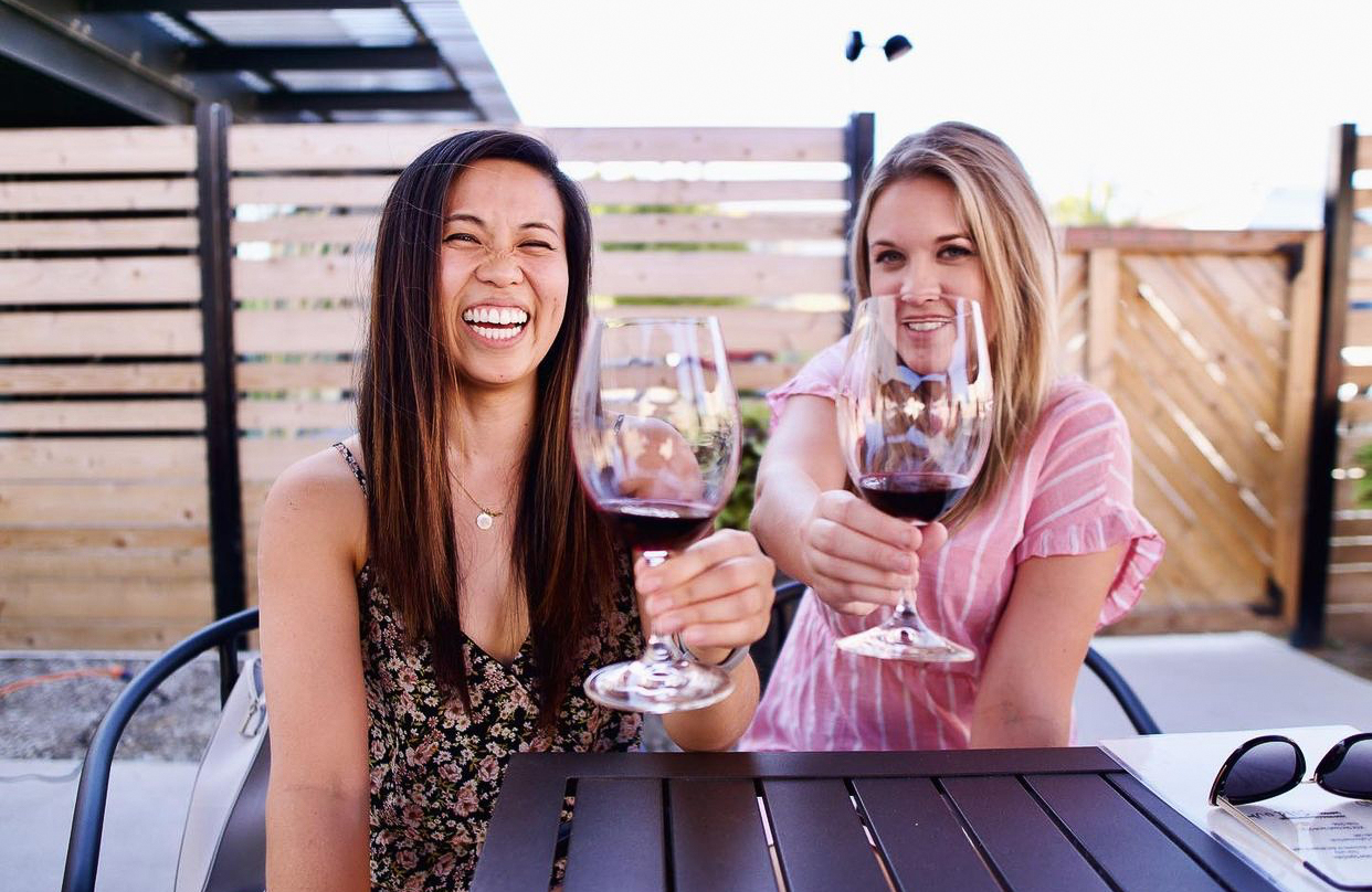 Two young woman smile and hold wine glasses with red wine up to the camera. The woman on the left has long, dark brown, straight hair in a black floral shirt and the other woman has medium length blonde hair in a striped pink shirt.