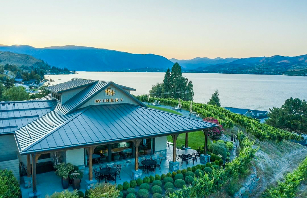 Building with overhanging roof and patio with tables and chairs in foreground surrounded by grape vines. A large lake and mountains are in the background, all in evening light.