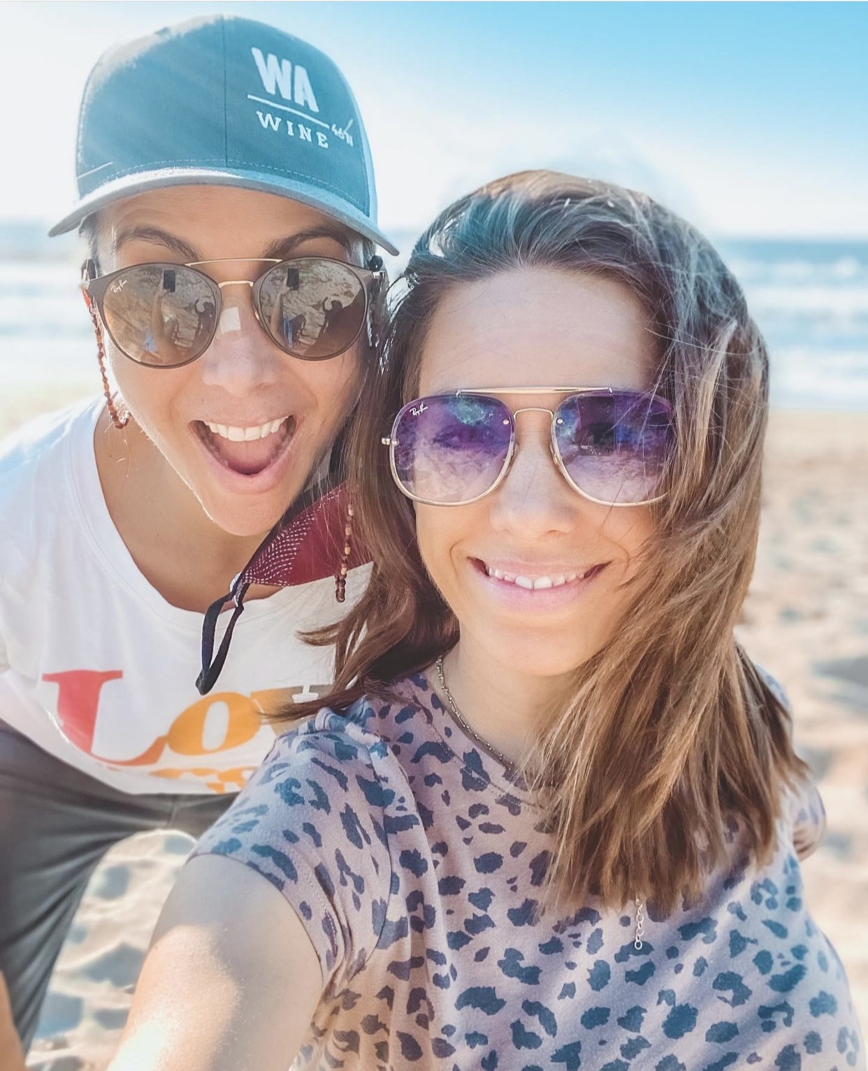 Two women in sunglasses smile in front of a sunny beach. The woman on the left wears a WA Wine ballcap.
