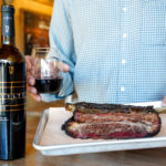 A man's torso is in frame. his left hand holds a plate with a large steak and his right hand holds a glass of red wine.