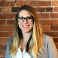 A young woman with long blond hair and dark glasses in a grey blazer smiles in front of a red brick wall