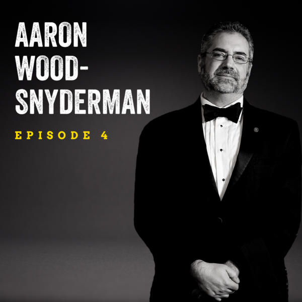Black and white image of a man standing in a black suit with bowtie smiling with his hands folded in front of him. Background is dark and the text overlay reads "Aaron Wood-Synderman Episode 4."