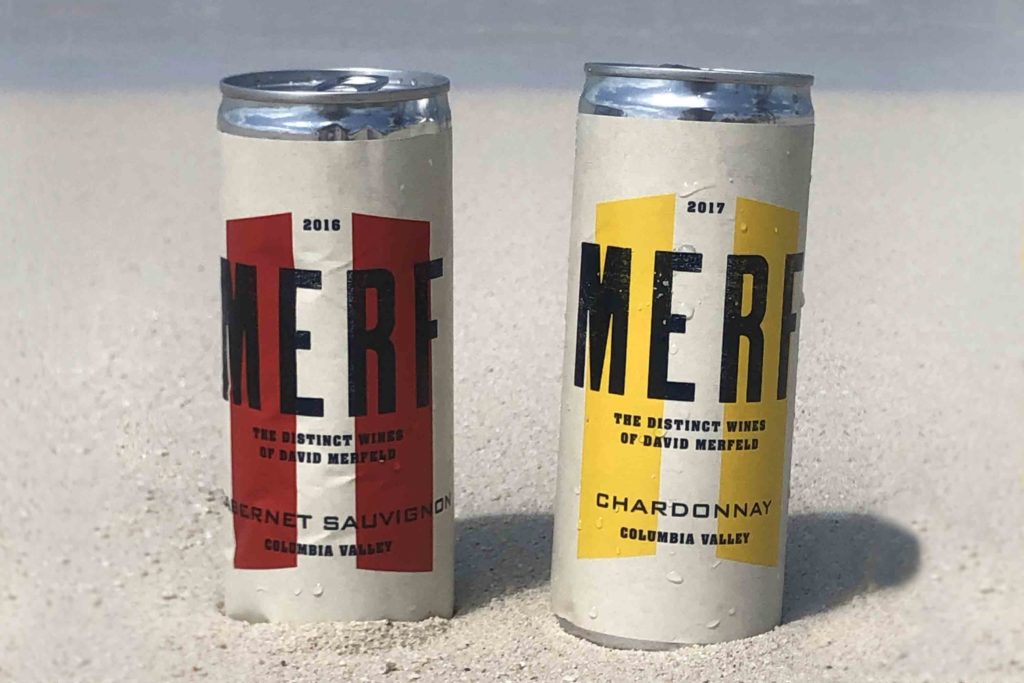 Two cans of wine, both with "Merf" on the label, one with red stripes and the other with yellow stripes. They are set in the sand of a beach.