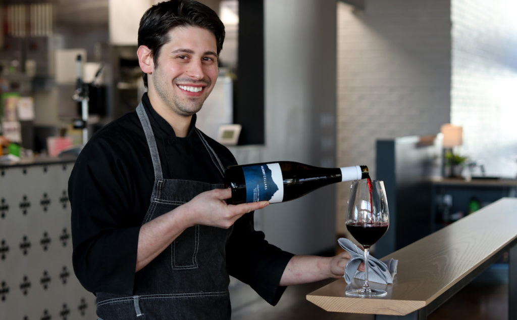 A man smiles at the camera as he pours red wine into a glass on a slender bar. A restaurant kitchen is out of focus behind him.