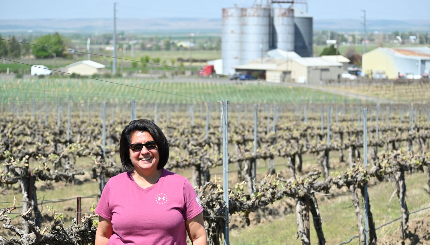 A woman in a pink shirt and sunglasses stands with short dark hair in front of vines without leaves, large cylindrical agricultural buildings are in the background