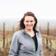 A woman with long dark hair in a grey rain jacket smiles in front of a bare vineyard.
