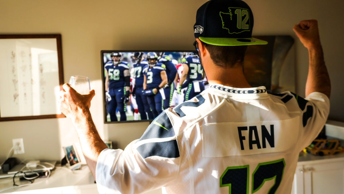A man holds a glass of red wine and is seen from the back wearing a "Fan" Seattle Seahawks white jersey and ball cap, watching a Seattle Seahawks football game.