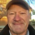 A man in a ballcap and black jacket smiles in front of sunlit building outdoors