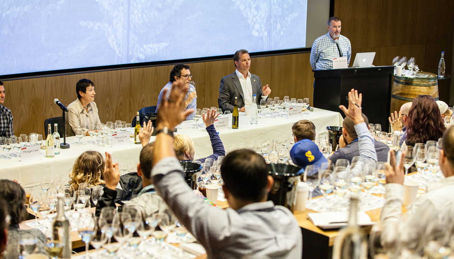 People sit at white tables with glasses of wine in front of them, raising their hands. There is a speaker at a podium and three other people seated at the front of the room.