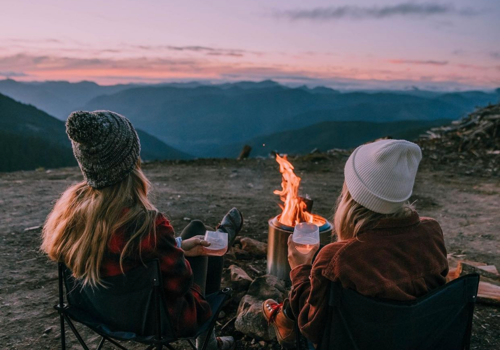View from behind of two women in beanies and sweaters sitting by a campfire with glasses of wine. They look out toward the mountains in the distance and the sky is faded pink and purple.