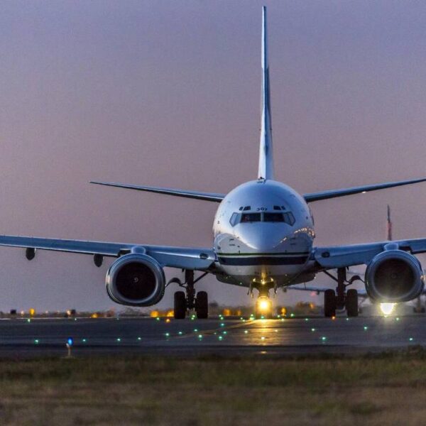 An airplane just above the tarmac in twilight. The plane's nose faces the viewer and a few bright lights shine from beyond the plane.