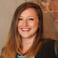 A woman with long red-brown hair in a black jacket and teal necklace smiles in front of a grey concrete wall