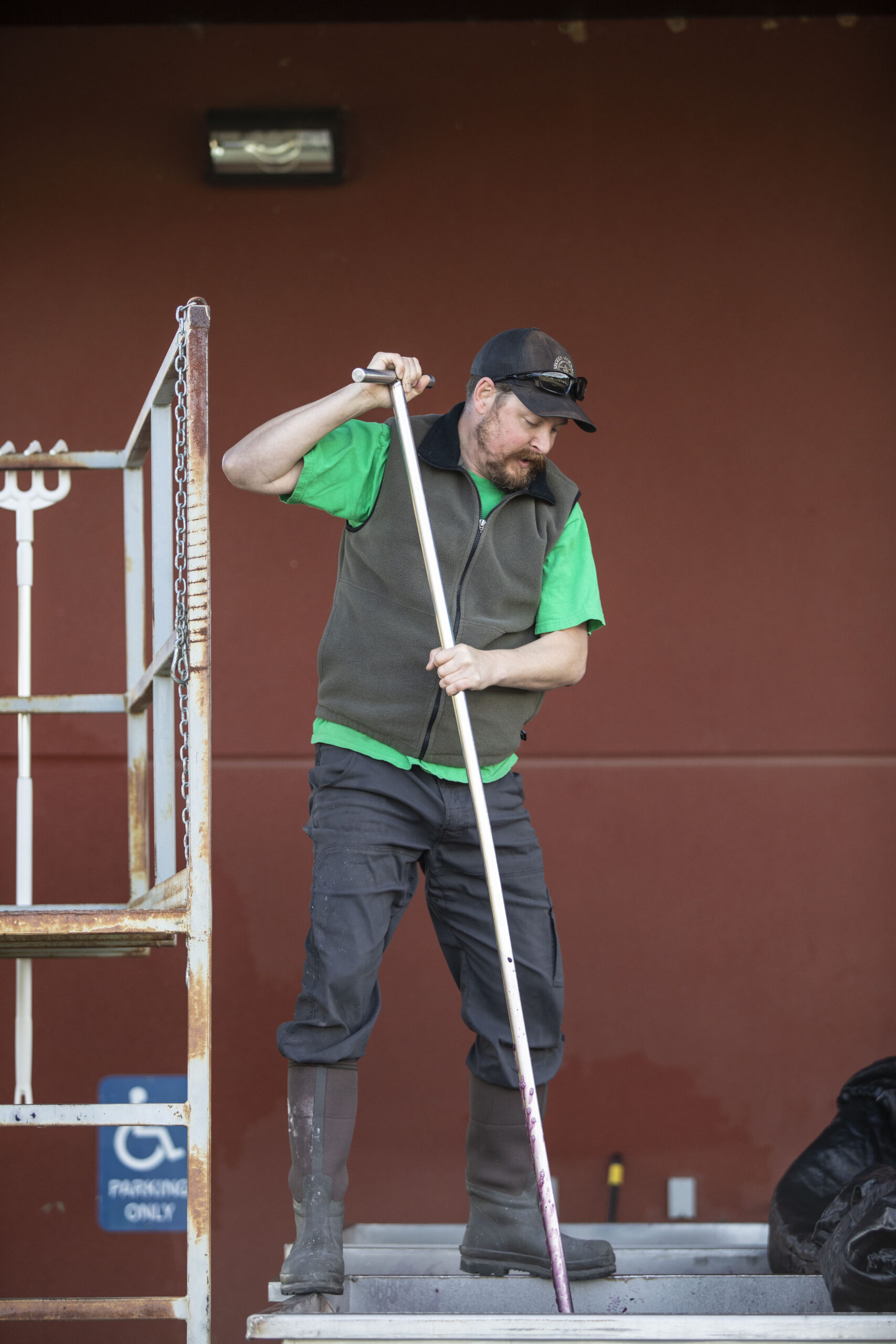 A man in a ballcap and grey vest stands on the edge of a metal container and pushes down a long metal pole into the bin. A red wall and metal structure are behind him.