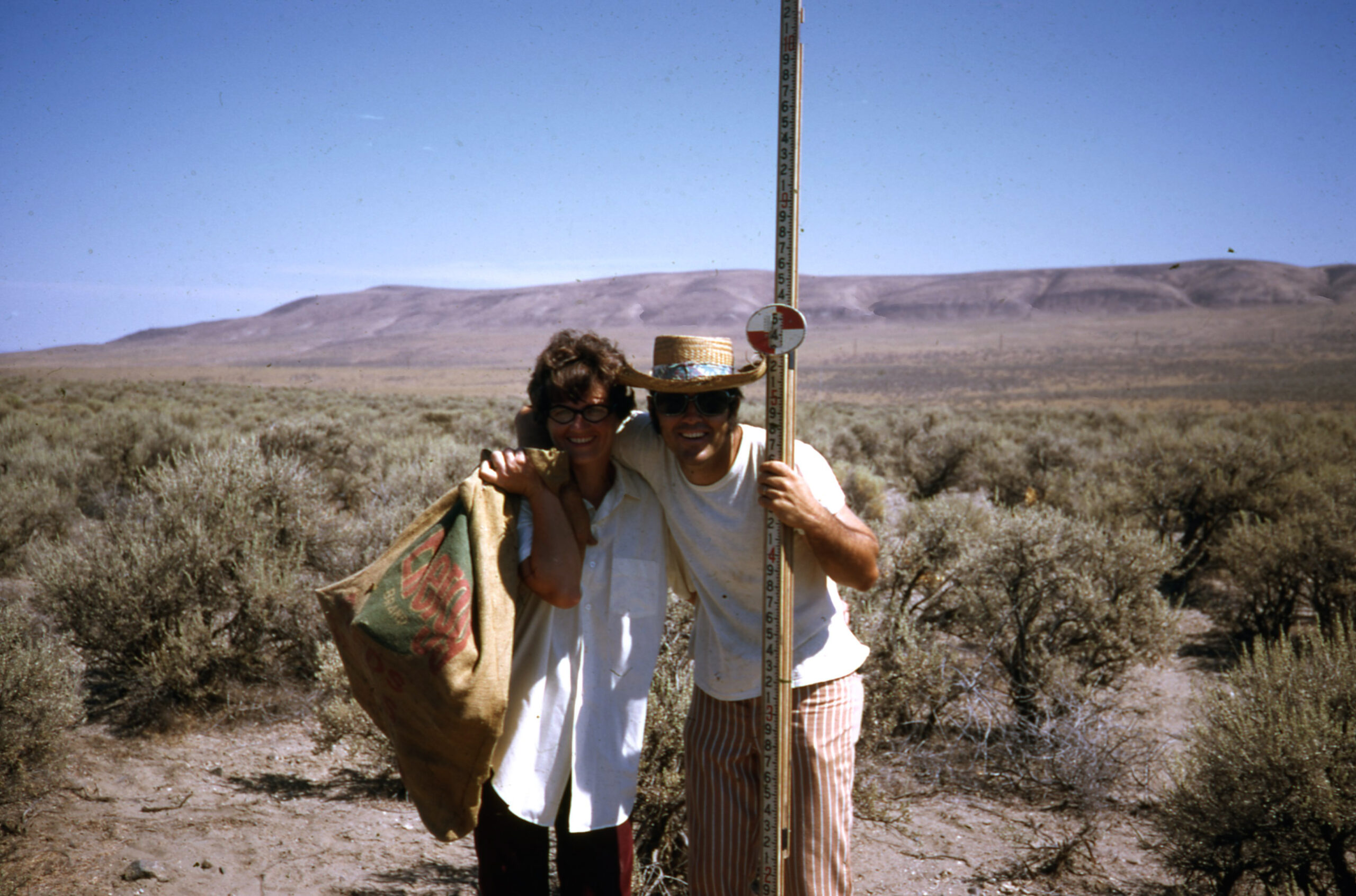 A man and a woman smile in a sagebrush field with rolling hills and blue sky behind. The woman holds a burlap sack over her shoulder and the man's arm is around her neck and holds a tall square measuring pole.