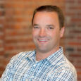 A man with short brown hair in a white and blue plaid shirt smiles in front of a red brick wall