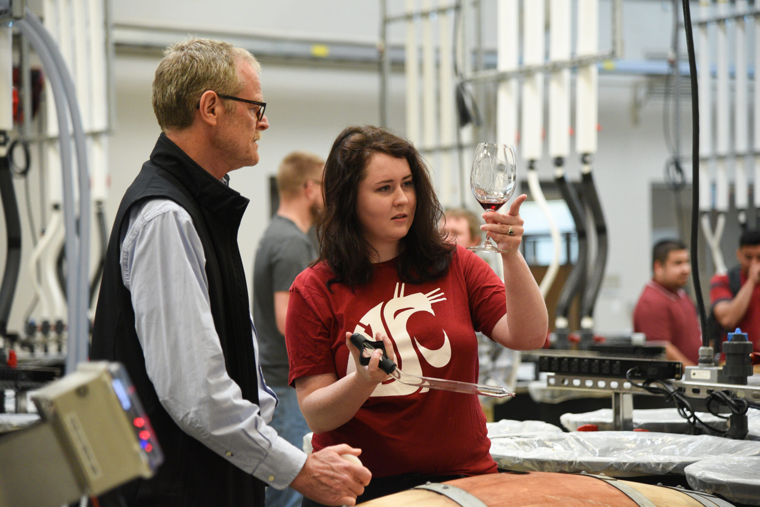 A young woman wearing a red WSU shirt holds a glass with red wine up to eye level and examines it. A man standing next to her wearing a black vest also looks at the glass. The background is a busy lab with tubes and other students.