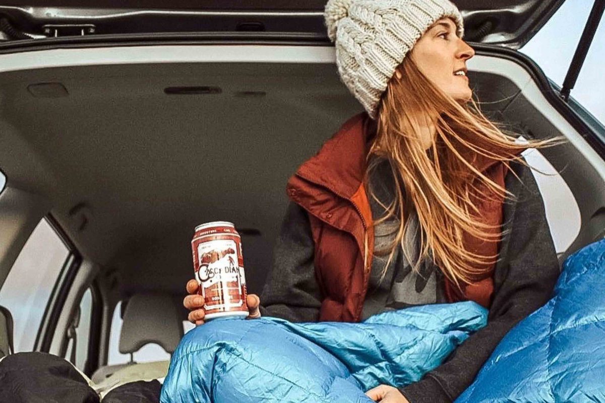 A woman with long auburn hair and a white beanie and red vest looks to her right with a can of wine in her left hand.