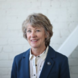 A woman with grey-brown hair in a blue blazer and white button-up shirt smiles in front of a white-washed brick wall