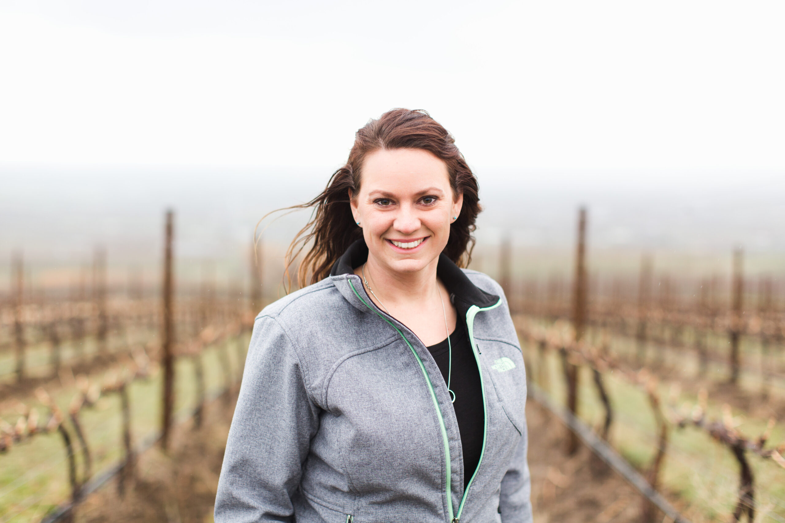 A woman with long dark hair and a grey jacket smiles and stand in front of grape vines without leaves, which are out of focus.