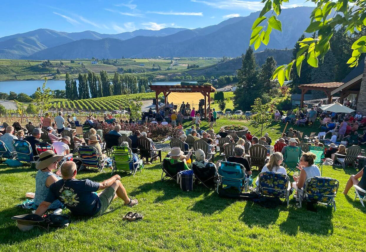 people sit in lawn chairs on bright green grass facing a stage with musicians, with green grape vines behind. Blue mountains are in the background.