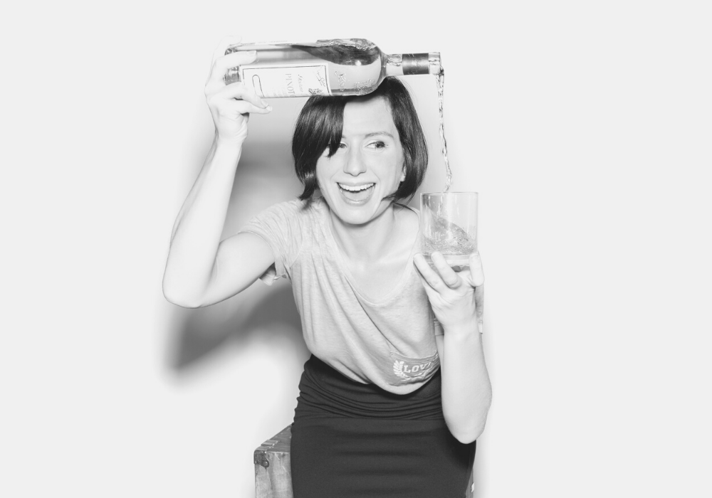 black and white image of woman with jaw-length dark hair pouring a liquid from a bottle into a glass in her other hand. She is bent forward and hold the bottle above her head.