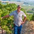 A man in a ballcap and plaid shirt smiles and stands next to a row of green vines.