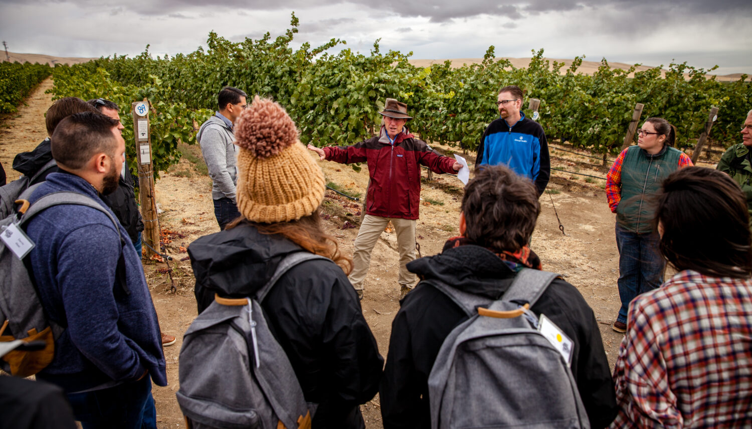 A man in a hat and red jacket is talking to a group of people in a circle. They stand in front of rows of grape vines and the sky is overcast.